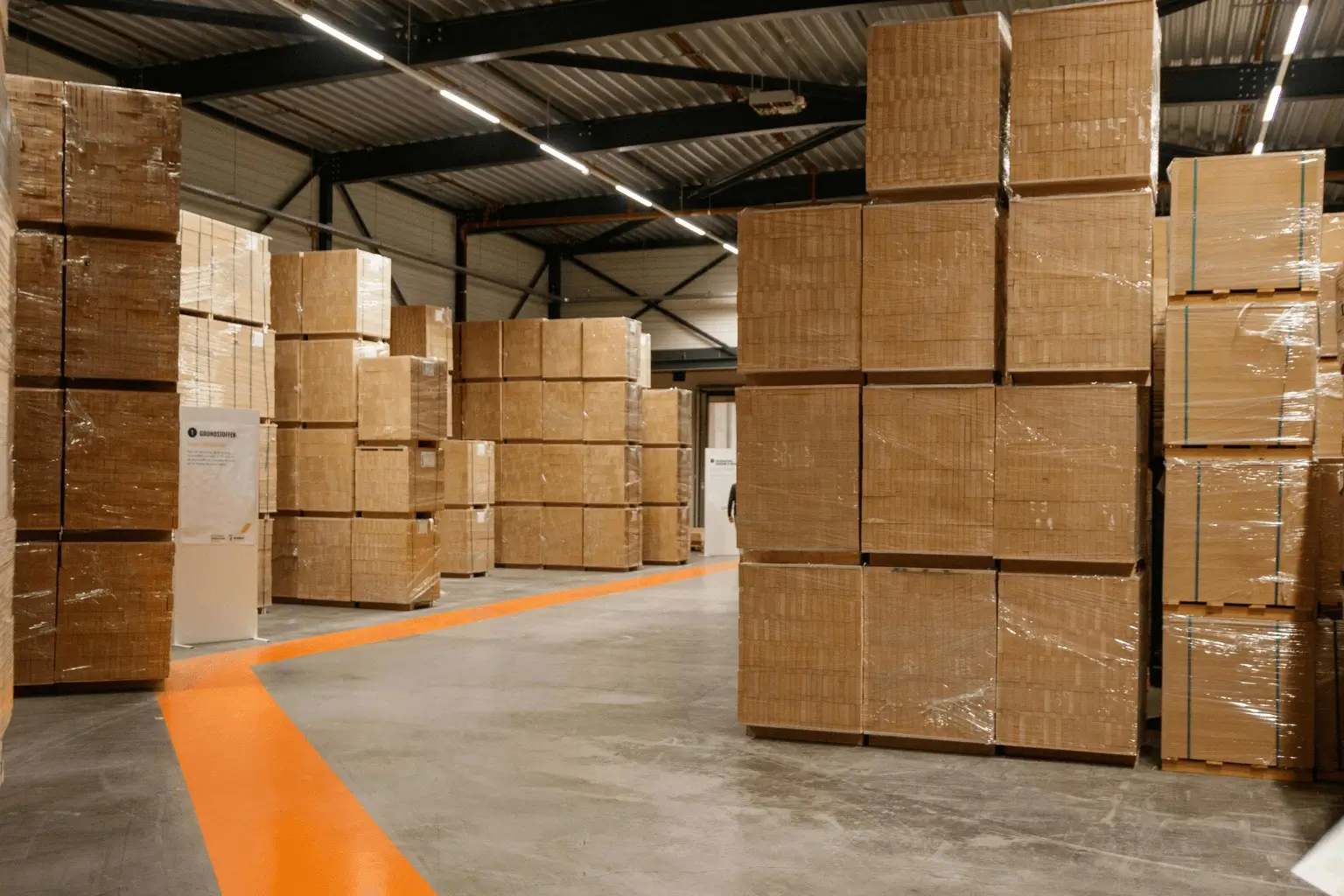 Magazyn materiałów budowlanych z wysokimi stosami produktów na paletach. Towary zabezpieczone folią i taśmami transportowymi. Przestrzeń magazynowa z wyznaczonym ciągiem komunikacyjnym.